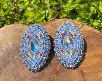 May include: A pair of light blue oval-shaped earrings with a beaded design. The earrings have a silver-colored border and a clear iridescent center.
