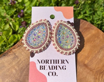 Puede incluir: Un par de pendientes en forma de lágrima iridiscentes con cuentas blancas y rosadas. Los pendientes están en una tarjeta blanca con el texto "NORTHERN BEADING CO."