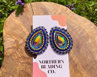 Puede incluir: Un par de pendientes en forma de lágrima con cuentas azules y doradas. Los pendientes presentan un gran cristal iridiscente en el centro. Los pendientes se muestran sobre una superficie de madera con una tarjeta que dice "Northern Beading Co."
