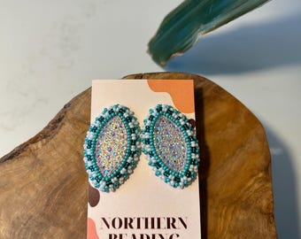 Puede incluir: Un par de pendientes ovalados con un diseño de cuentas de color verde azulado y blanco. Los pendientes están en una tarjeta que dice "Northern Beading Co."