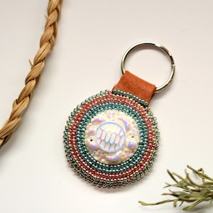 May include: A beaded keychain with a round, multi-coloured design featuring a white iridescent turtle in the centre. The keychain is attached to a brown leather strap and a silver ring.