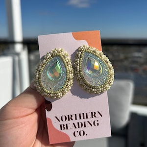 Puede incluir: Un par de pendientes de cuentas verdes y blancas con piedras iridiscentes en forma de lágrima. Los pendientes están en una tarjeta blanca con el texto "NORTHERN BEADING CO."