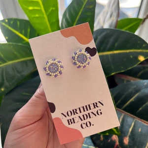 Peut inclure: Une paire de petites boucles d'oreilles rondes en perles avec un centre blanc et un anneau extérieur violet. Les boucles d'oreilles sont présentées sur une carte avec le texte "Northern Beading Co."