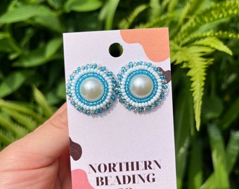 Puede incluir: Un par de pendientes de cuentas con un centro de perla blanca y cuentas azules y blancas. Los pendientes están en una tarjeta blanca con el texto "NORTHERN BEADING CO."