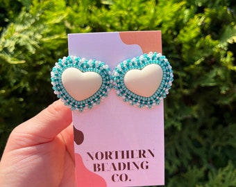 Puede incluir: Un par de pendientes en forma de corazón con un centro blanco y un borde de cuentas de color turquesa y blanco. Los pendientes se muestran en una tarjeta con el texto "Northern Beading Co."