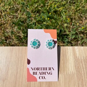 Puede incluir: Un par de peque&ntilde;os pendientes de cuentas blancas con un centro turquesa. Los pendientes se muestran en una tarjeta blanca con el texto "NORTHERN BEADING CO."