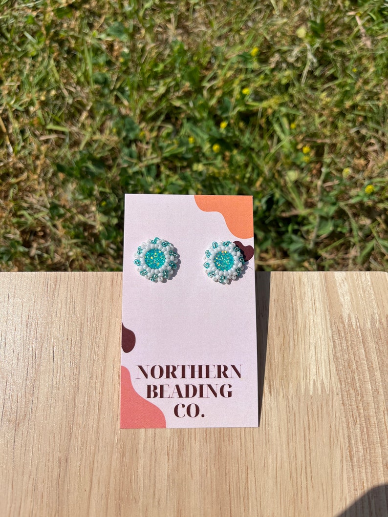 Puede incluir: Un par de peque&ntilde;os pendientes de cuentas blancas con un centro turquesa. Los pendientes se muestran en una tarjeta blanca con el texto "NORTHERN BEADING CO."