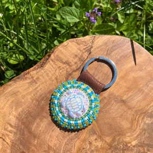 Può includere: Un portachiavi a forma di tartaruga in perline con un designo di tartaruga bianco iridescente, circondato da perline blu e verdi. Il portachiavi è attaccato a un cinturino in pelle marrone e un anello portachiavi argentato.