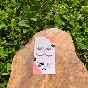 Può includere: Orecchini a cerchio in argento e perle bianche con un ciondolo a forma di fiore bianco. Gli orecchini sono esposti su una superficie in legno con un biglietto che recita "Northern Beading Co."