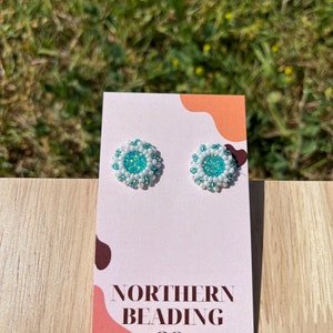 Puede incluir: Un par de peque&ntilde;os pendientes redondos con un dise&ntilde;o de cuentas blancas y turquesa. Los pendientes se muestran en una tarjeta blanca con el texto "NORTHERN BEADING CO."