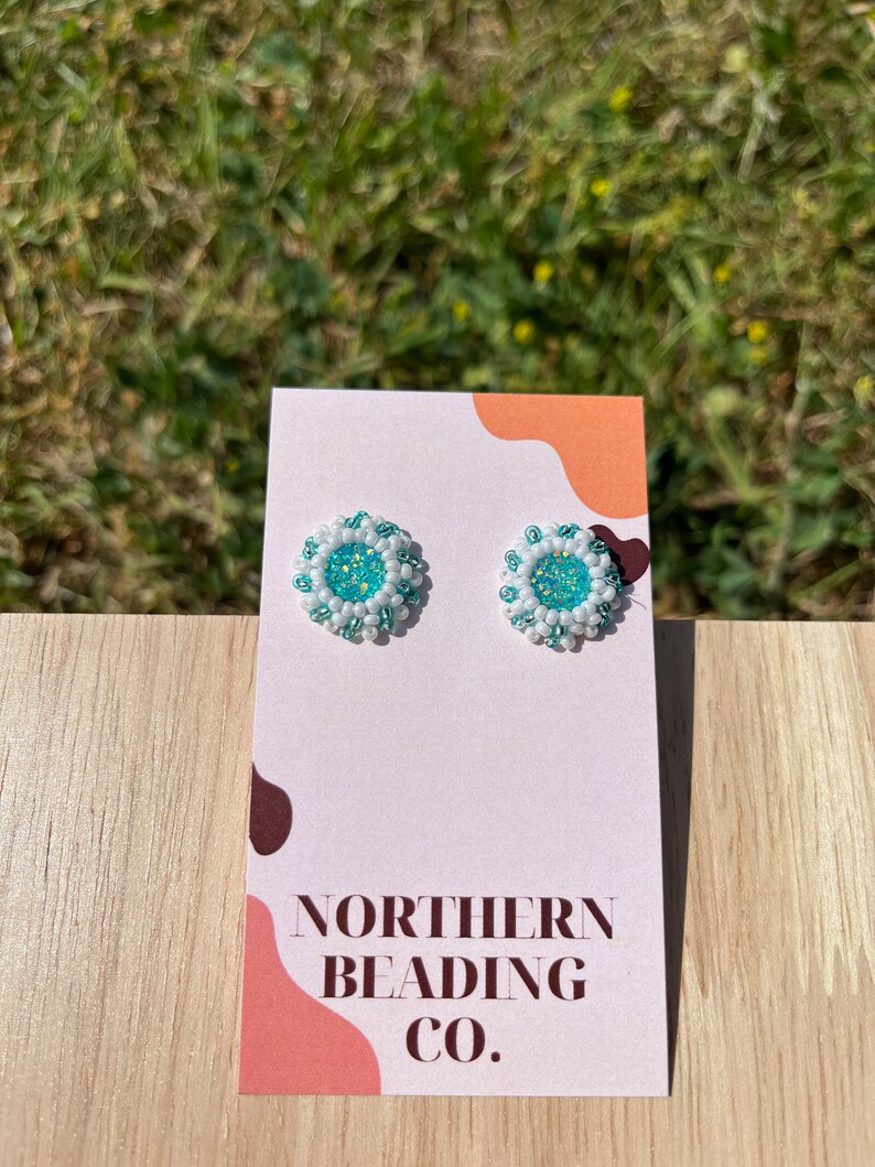 Puede incluir: Un par de peque&ntilde;os pendientes redondos con un dise&ntilde;o de cuentas blancas y turquesa. Los pendientes se muestran en una tarjeta blanca con el texto "NORTHERN BEADING CO."