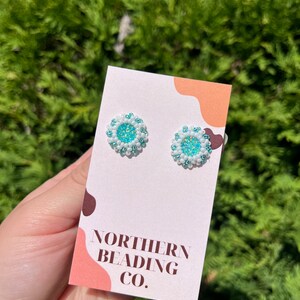 Puede incluir: Un par de peque&ntilde;os pendientes redondos con un centro turquesa rodeado de cuentas blancas. Los pendientes se muestran en una tarjeta blanca con el texto "NORTHERN BEADING CO."