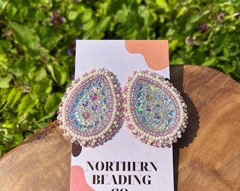 Puede incluir: Un par de pendientes de cuentas rosas y blancas con detalles iridiscentes. Los pendientes tienen forma de lágrima y un borde de cuentas blancas. Los pendientes se muestran en una tarjeta blanca con el texto "NORTHERN BEADING CO."