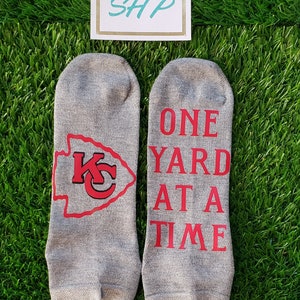 May include: A pair of gray ankle socks with red lettering. One sock has the Kansas City Chiefs logo and the other says "One Yard At A Time".