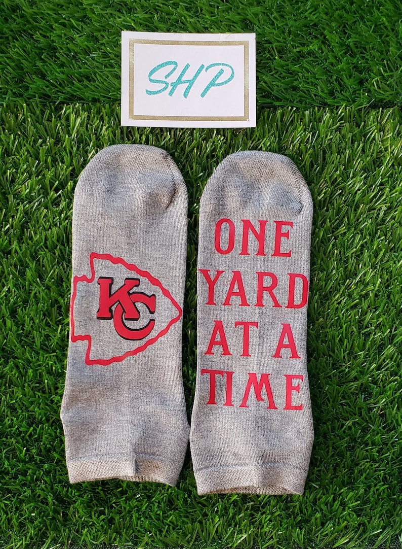 May include: A pair of gray ankle socks with red lettering. One sock has the Kansas City Chiefs logo and the other says "One Yard At A Time".