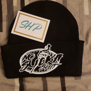 Op de afbeelding: Zwarte beanie met een wit ovaal logo met de tekst "No Sucka Shit" en een kleine afbeelding van een schedel met een kroon. De beanie heeft een wit label met de tekst "SHP" erop.