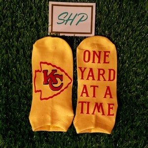 May include: A pair of yellow ankle socks with red and white lettering. One sock has a red and white Kansas City Chiefs logo. The other sock says "ONE YARD AT A TIME".