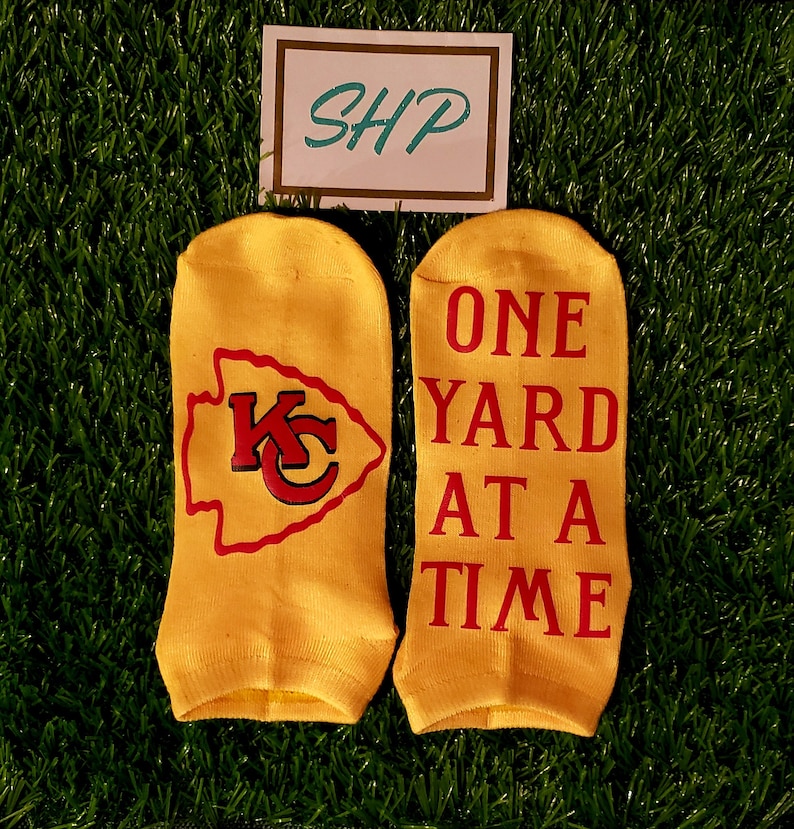 May include: A pair of yellow ankle socks with red and white lettering. One sock has a red and white Kansas City Chiefs logo. The other sock says "ONE YARD AT A TIME".