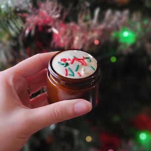 May include: A small, brown jar with a white topping and colourful sprinkles. The jar is held in a hand, with a blurred Christmas tree in the background. The sprinkles are red and green.