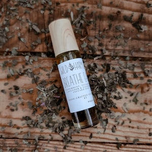 May include: A clear glass essential oil roller bottle with a wooden cap. The label reads "WILD HARMONY BREATHE EASY" with ingredients listed. The bottle is on a wooden surface scattered with dried herbs.