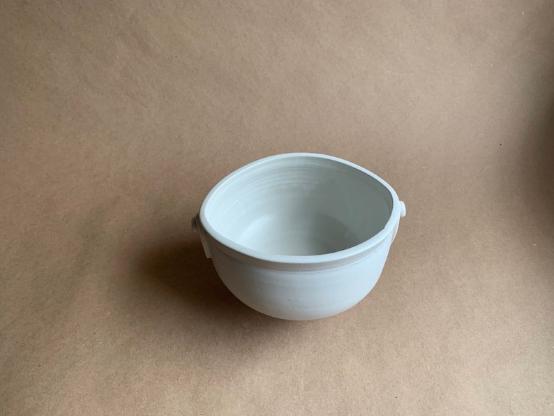 May include: A white ceramic bowl with two small handles on the sides. The bowl is round and has a smooth, glossy finish.