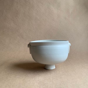 May include: A white ceramic bowl with two small handles on the sides and a pedestal base.