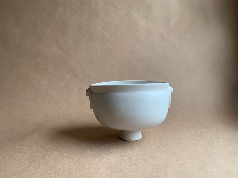 May include: A white ceramic bowl with two small handles on the sides and a pedestal base.