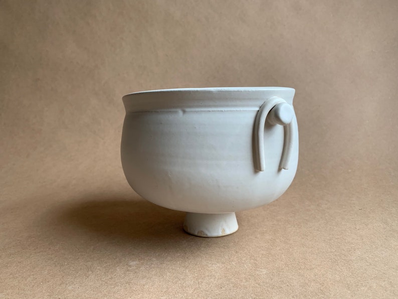 May include: White ceramic planter with a rounded body and a single handle. The handle is a loop with a small knob on the end. The planter sits on a short, round pedestal.