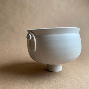May include: White ceramic bowl with a rounded base and a single handle on one side. The bowl has a smooth, matte finish.