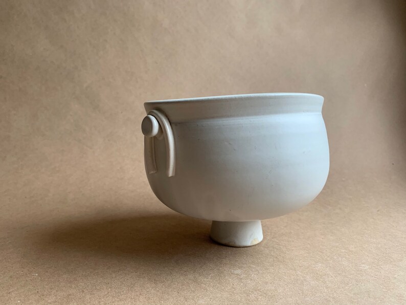 May include: White ceramic bowl with a rounded base and a single handle on one side. The bowl has a smooth, matte finish.