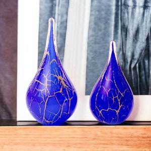 Op de afbeelding: Twee blauwe glazen vazen in de vorm van een traan met gouden craquelé-patronen. De vazen staan op een houten oppervlak.