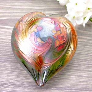 Puede incluir: Un pisapapeles de vidrio en forma de corazón con un patrón de remolino de rosa, naranja y verde. El vidrio es iridiscente y refleja la luz.