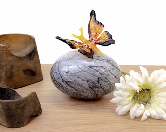 May include: A glass butterfly figurine with orange, yellow, and purple wings perched on a gray marble orb. A white gerbera daisy sits on a wooden surface.