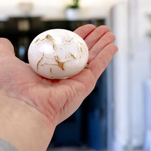 Puede incluir: Una esfera blanca con vetas doradas, sostenida en una mano.