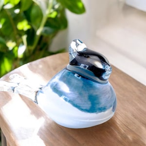 Peut inclure: Figurine d'oiseau en verre avec un corps bleu et blanc et une tête noire. L'oiseau a les ailes déployées et est posé sur une surface en bois. La figurine est un objet décoratif.
