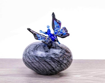 May include: A blue glass butterfly figurine perched atop a gray marble orb. The butterfly has iridescent wings and a detailed body.