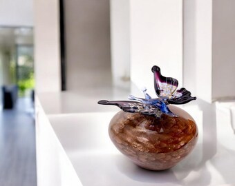 May include: A glass butterfly sculpture with purple, blue, and gold accents sits on a brown, round, textured base.