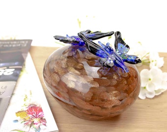 May include: A glass orb with a gold and brown swirl pattern, topped with two blue glass butterflies. The butterflies have black accents and are positioned on top of the orb.