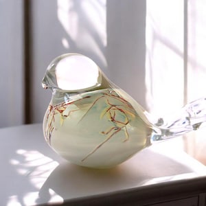 Könnte beinhalten: Eine klare Glasvogelfigur mit einem weißen, gelben und roten Wirbelmuster. Der Vogel sitzt auf einer weißen Oberfläche.