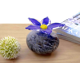 May include: A glass flower with blue petals and a yellow center sits on top of a round, gray and black glass vase. The vase is sitting on a wooden surface.