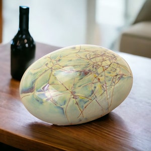 Könnte beinhalten: Eine große, hellgrüne Keramikvase mit einer craquelé-Glasur und einem wirbelnden Muster aus braunen und blauen Linien. Die Vase steht auf einem Holztisch.