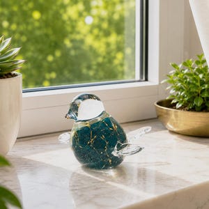 Könnte beinhalten: Eine Glasvogel-Figur mit einem tief türkisfarbenen Körper, der mit goldenen Sprenkeln und Adern durchzogen ist. Der Kopf besteht aus klarem Glas mit einem weißen, undurchsichtigen Fleck, während Flügel und Schwanz aus glattem, klarem Glas sind.