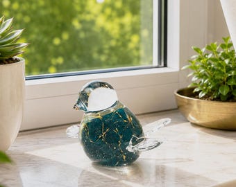 May include: A glass bird figurine featuring a deep teal body with gold-colored flecks and veining throughout. The head is clear glass with a white opaque patch, and the wings and tail are made of clear, smooth glass, creating a reflective surface.