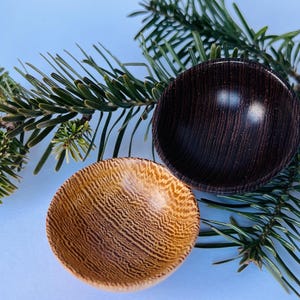 Op de afbeelding: Twee kleine houten kommen, één licht en één donker, met een natuurlijk houtnerfpatroon, rusten op een bedje van groene dennennaalden. De kommen zijn rond en lijken met de hand gedraaid te zijn, waarbij de textuur en kleurvariaties van het hout worden benadrukt.