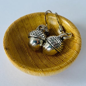 May include: Silver acorn earrings with detailed caps sit in a small, round, wooden dish. The earrings have a textured design on the caps and a smooth, rounded body. The dish is light brown with a natural wood grain.