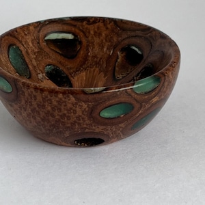 Op de afbeelding: Een kleine, handgemaakte houten kom met een uniek ontwerp. De kom is gemaakt van donkerbruin hout met natuurlijke patronen en ingelegd met groene en zwarte accenten. De kom heeft een gladde, gepolijste afwerking en is ideaal voor het bewaren van kleine items.