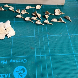 Puede incluir: Una colecci&oacute;n de peque&ntilde;as formas de papel troquelado de color beige claro que se asemejan a p&eacute;talos de flores u hojas. Algunas piezas est&aacute;n dispuestas en un grupo suelto a la izquierda, mientras que otras est&aacute;n esparcidas individualmente.