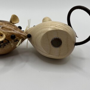 Puede incluir: Dos figuras de madera con forma de rat&oacute;n y cuerpos lisos y redondeados. Un rat&oacute;n presenta una veta de madera oscura y veteada con ojos de cuentas negras y orejas claras. El otro es de madera clara con un inserto magn&eacute;tico circular y una cola negra.