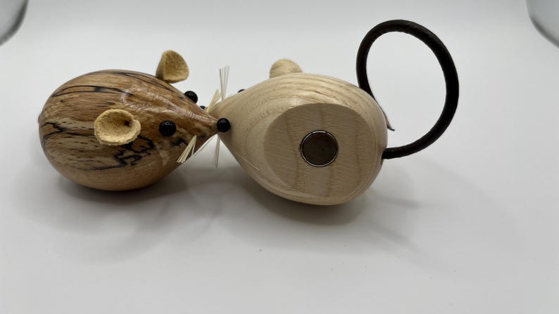 Puede incluir: Dos figuras de madera con forma de rat&oacute;n y cuerpos lisos y redondeados. Un rat&oacute;n presenta una veta de madera oscura y veteada con ojos de cuentas negras y orejas claras. El otro es de madera clara con un inserto magn&eacute;tico circular y una cola negra.