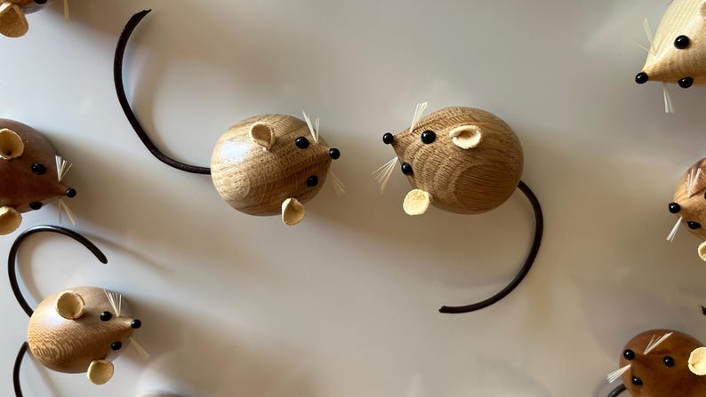 Puede incluir: Varias figuras peque&ntilde;as de ratones de madera redondeadas dispuestas en un patr&oacute;n. Cada rat&oacute;n presenta un cuerpo de madera de color marr&oacute;n claro con vetas, dos orejas peque&ntilde;as redondeadas, ojos de cuentas negras, un hocico puntiagudo con bigotes blancos finos y una cola larga, fina y oscura.